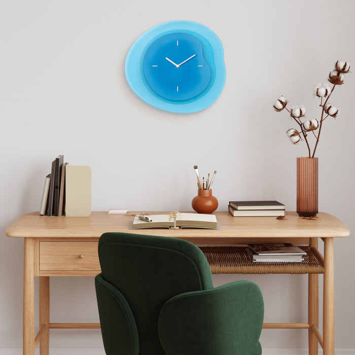 Lifestyle image of the 404-3039BL 15.2-inch Drift Wall Clock – Blue, shown hanging on a cream wall in a Transitional Style home office with a light wood desk and green velvet chair. The clock features an open face multi-layered design with drifting contours, metal hands, and raised number markings in white.