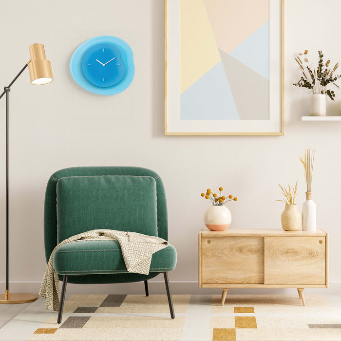 Lifestyle image of the 404-3039BL 15.2-inch Drift Wall Clock – Blue, shown hanging on a tan wall above an emerald green chenille chair, retro brass floor lamp, and light wood side cabinet. The clock features an open face multi-layered design with drifting contours, metal hands, and raised number markings in white.