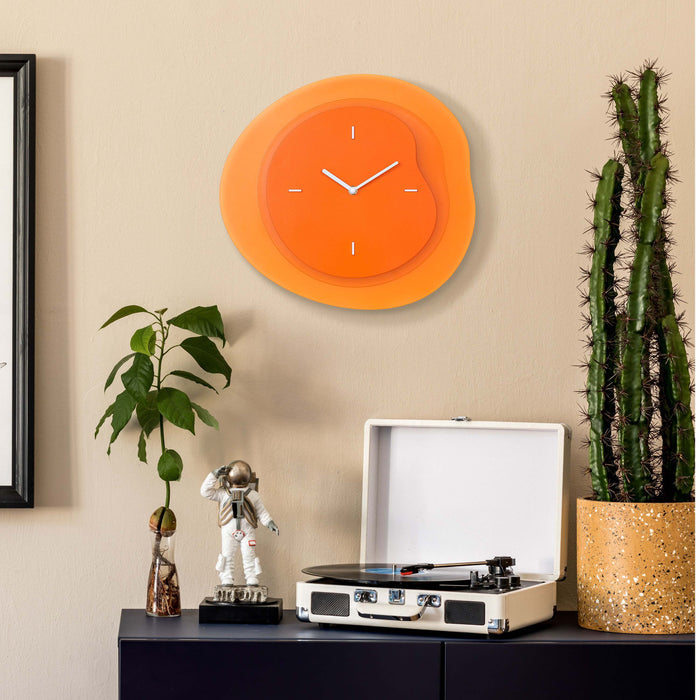 Lifestyle image of the 404-3039O 15.2-inch Drift Wall Clock – Orange, shown hanging on a tan wall above a black side table with a vintage turntable, a cactus, a plant, and a statue of an astronaut. The clock features an open face multi-layered design with drifting contours, metal hands, and raised number markings in white.