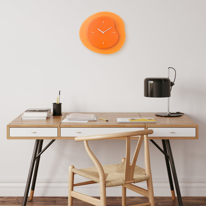 Lifestyle image of the 404-3039O 15.2-inch Drift Wall Clock – Orange, shown hanging on a light wall above a modern black and natural wood desk. The clock features an open face multi-layered design with drifting contours, metal hands, and raised number markings in white.