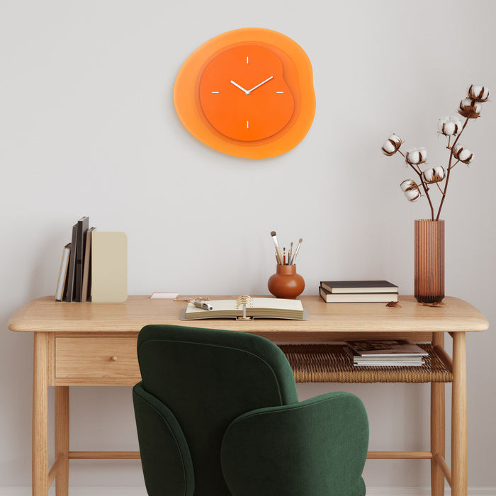 Lifestyle image of the 404-3039O 15.2-inch Drift Wall Clock – Orange, shown hanging on a cream wall in a Transitional Style home office with a light wood desk and green velvet chair. The clock features an open face multi-layered design with drifting contours, metal hands, and raised number markings in white.