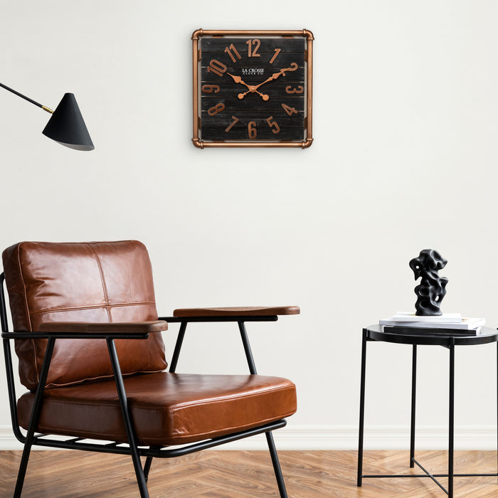 Lifestyle image of the 404-3412D 16-inch Foundry Metal & Wood Wall Clock - Dark, shown hanging on a white wall above a cognac leather chair and side table. The clock features a square metal pipe frame in a copper finish, a black-stained wood slat clock face with copper-finished metal numbers and hands.