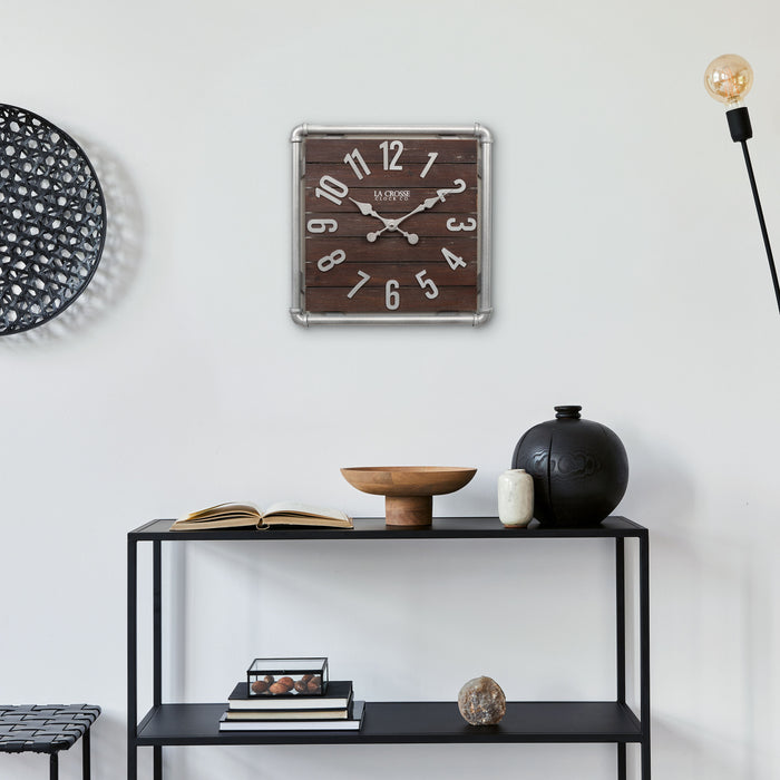 Lifestyle image of the 404-3412L 16-inch Foundry Metal & Wood Wall Clock - Light, shown hanging above a black metal shelving unit with books and vases. The clock features a square metal pipe frame in a weathered pewter finish, a truffle-brown-stained wood slat clock face with pewter-finished metal numbers and hands.