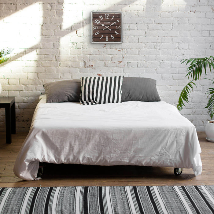 Lifestyle image of the 404-3412L 16-inch Foundry Metal & Wood Wall Clock - Light, shown hanging on a white-washed brick wall above a bed. The clock features a square metal pipe frame in a weathered pewter finish, a truffle-brown-stained wood slat clock face with pewter-finished metal numbers and hands.