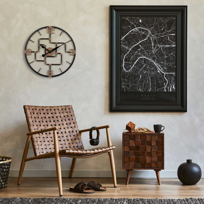 Lifestyle image of the 404-3420 20-inch Pipeworks Metal Wall Clock shown hanging on a tan plaster finish wall hanging next to map artwork, and above a dark wicker chair with a woven wood side table. The clock features an open face dial with a weathered bronze and antique brass finish pipe framework, and antique brass numbers 3, 6, 9, and 12.