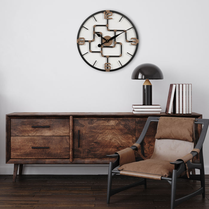 Lifestyle image of the 404-3420 20-inch Pipeworks Metal Wall Clock shown hanging on a white wall above a Mid-Century Modern console table with a retro black lamp and a brown architectural chair. The clock features an open face dial with a weathered bronze and antique brass finish pipe framework, and antique brass numbers 3, 6, 9, and 12.
