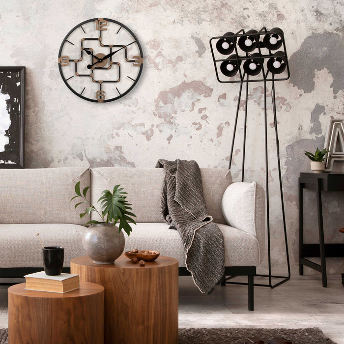 Lifestyle image of the 404-3420 20-inch Pipeworks Metal Wall Clock shown hanging on a distressed concrete wall above a tan modern couch with cylindrical wood tables, and a floor lamp. The clock features an open face dial with a weathered bronze and antique brass finish pipe framework, and antique brass numbers 3, 6, 9, and 12.