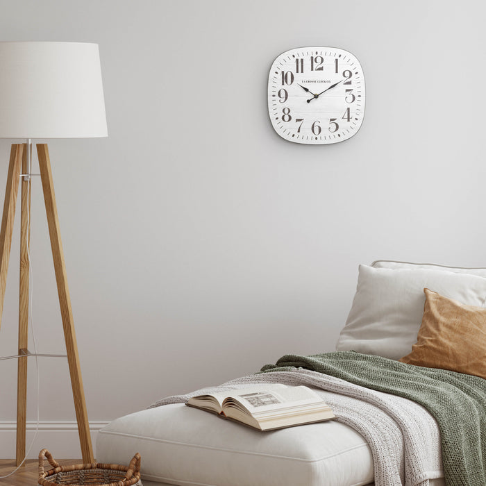 404-3428 11-inch Sylvie square MDF open face wall clock featuring a faux whitewash wood dial black numbers and metal hands hanging on a white wall over a chaise lounge with a wood floor lamp