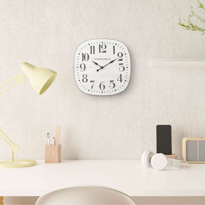 404-3428 11-inch Sylvie square MDF open face wall clock featuring a faux whitewash wood dial black numbers and metal hands hanging on a beige textured wall over a neutral colored desk with neutral accents