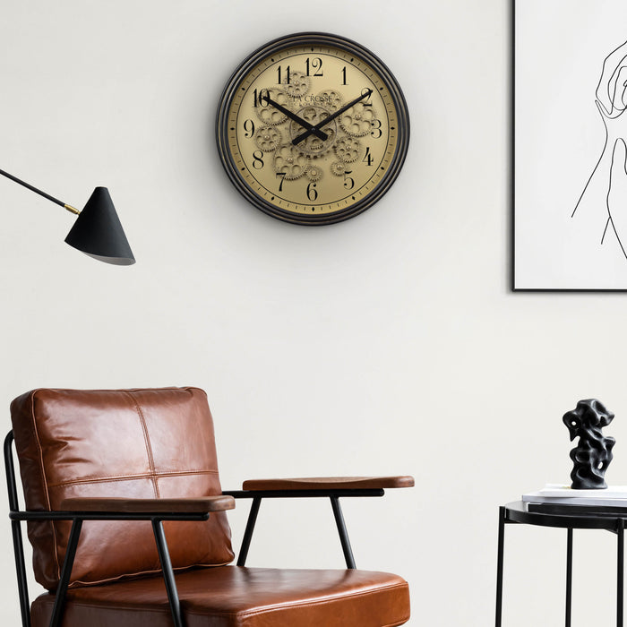 Lifestyle image of the 404-3439 15-inch Moving Gears Wall Clock hanging on a wall above a leather chair and side table with a masculine modern decor. The clock features a metal oil-rubbed antique frame, distressed brushed dial with moving gears, and black numbers and hands.