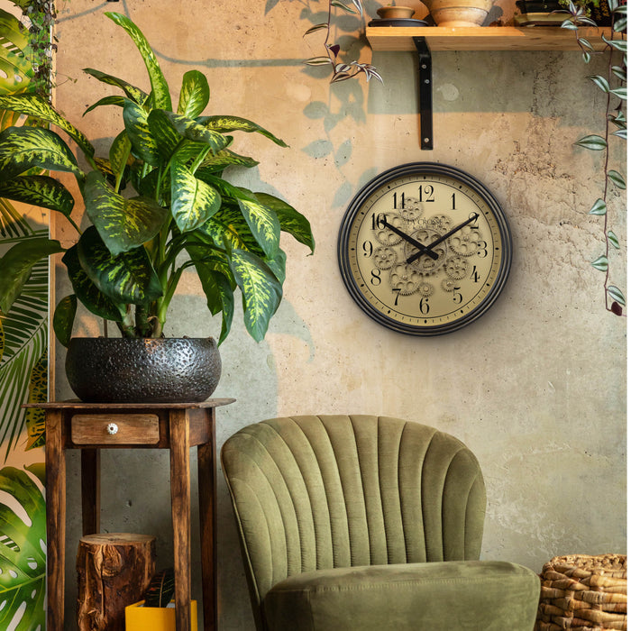 Lifestyle image of the 404-3439 15-inch Moving Gears Wall Clock hanging on a tan textured wall above a green velvet chair and wood plant stand. The clock features a metal oil-rubbed antique frame, distressed brushed dial with moving gears, and black numbers and hands.