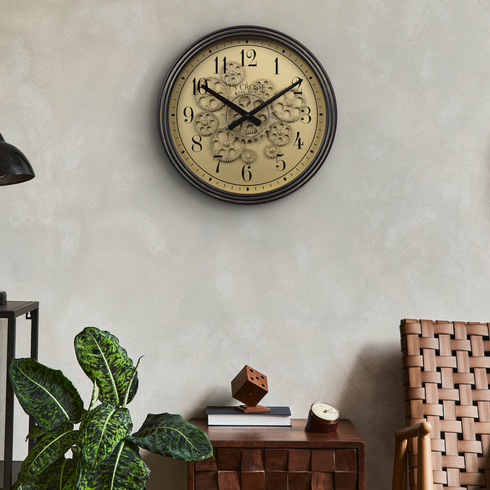 Lifestyle image of the 404-3439 15-inch Moving Gears Wall Clock hanging on a wall above a chair and side table with a tropical theme decor. The clock features a metal oil-rubbed antique frame, distressed brushed dial with moving gears, and black numbers and hands.