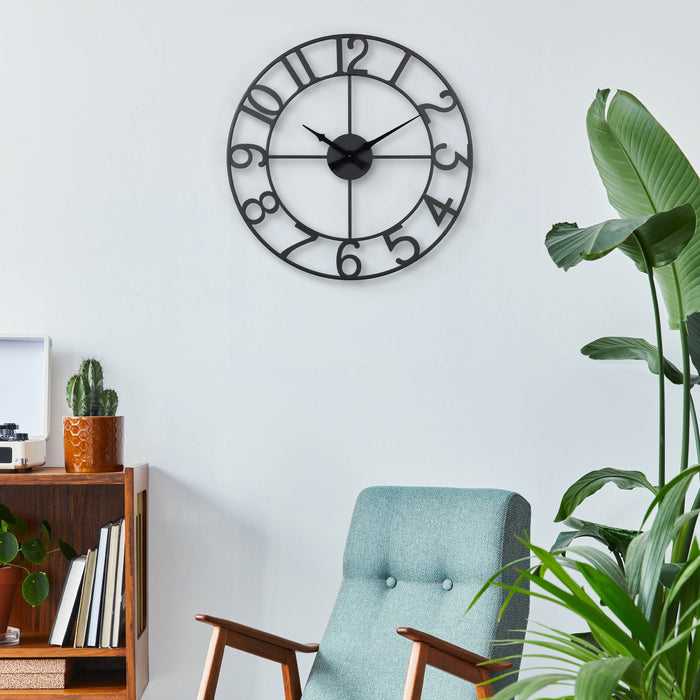 Lifestyle image of the 404-3451A 20-inch Metal Tower Wall Clock - Arabic hanging on a light blue wall over a mid-century modern chair. This skeleton tower clock features Arabic numbers, hour and minute hands all constructed in metal.