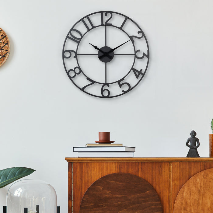 Lifestyle image of the 404-3451A 20-inch Metal Tower Wall Clock - Arabic hanging on a wall above wood side table. This skeleton tower clock features Arabic numbers, hour and minute hands all constructed in metal.