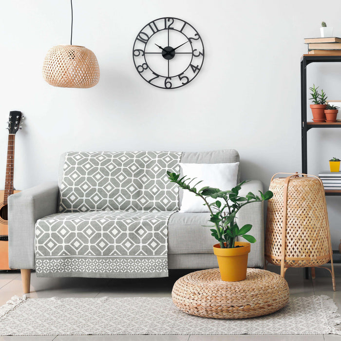 Lifestyle image of the 404-3451A 20-inch Metal Tower Wall Clock - Arabic hanging on a wall above a gray couch with Bohemian accents, This skeleton tower clock features Arabic numbers, hour and minute hands all constructed in metal.