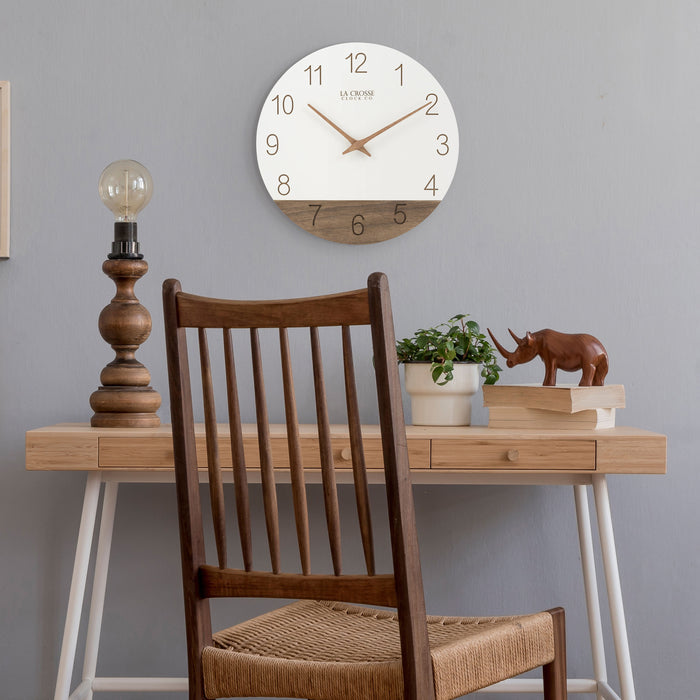 Lifestyle image of the 404-3630B 12-inch Sierra Wood Wall - White, hanging on a gray wall above a Scandinavian style desk of white and light wood with a lamp, a potted plant, a wood rhinocerous sculpture and a Transitional style wood chair. The clock features laser-cut numbers, a black and wood open-face dial, and wood hour and minute hands.