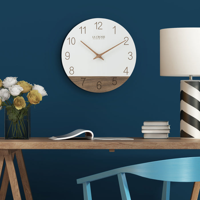 Lifestyle image of the 404-3630B 12-inch Sierra Wood Wall - White, hanging on a dark royal blue wall above a mid-tone wood desk with yellow and white roses in a vase, a stack of books, a black and white geometric lamp, and a light blue wood chair. The clock features laser-cut numbers, a black and wood open-face dial, and wood hour and minute hands.