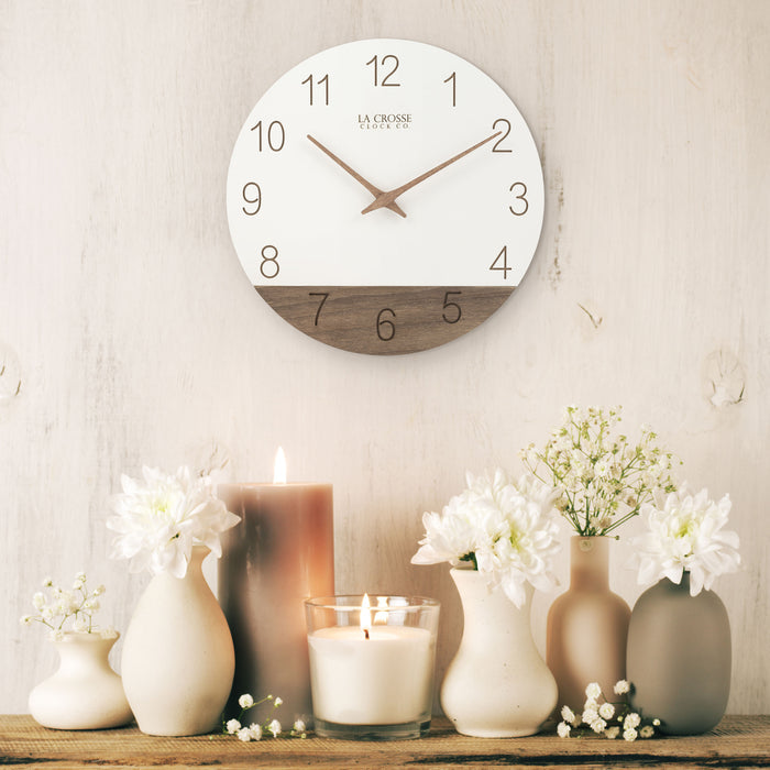 Lifestyle image of the 404-3630B 12-inch Sierra Wood Wall - White, hanging on a cream paneled wall above a wooden rustic shelf holding vases of flowers and candles. The clock features laser-cut numbers, a black and wood open-face dial, and wood hour and minute hands.