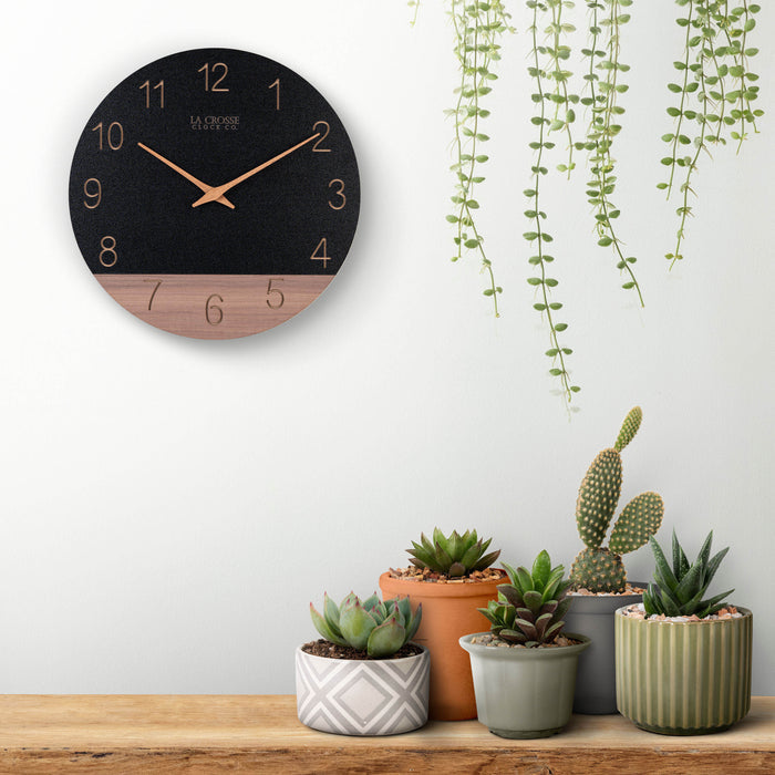 Lifestyle image of the 404-3630C 12-inch Sierra Wood Wall Clock - Black, hanging on a wall surrounded by plants. The clock features a black and wood split design open-face dial, with laser cut numbers, and wood hour and minute hands.