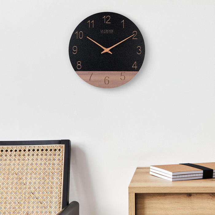 Lifestyle image of the 404-3630C 12-inch Sierra Wood Wall Clock - Black, the clock is shown hanging on a wall above a side table and a wicker chair. The clock features a black and wood split design open-face dial, with laser cut numbers, and wood hour and minute hands.