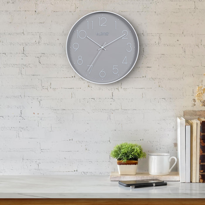 Lifestyle image of the 404-3831 12-inch Paxton Wall Clock - Gray, with Silent Sweep, hanging on a white-washed brick wall above a marble countertop with books, a plant, and a phone. The clock features a flush-mount gray frame, a matching color dial, white raised numbers, and white metal hour, minute, and second hands.