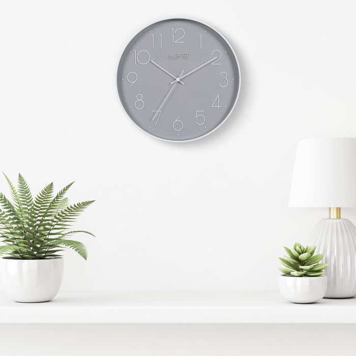 Lifestyle image of the 404-3831 12-inch Paxton Wall Clock - Gray, with Silent Sweep, hanging on a white wall above a white shelf with a white lamp and plants in white vases. The clock features a flush-mount gray frame, a matching color dial, white raised numbers, and white metal hour, minute, and second hands.