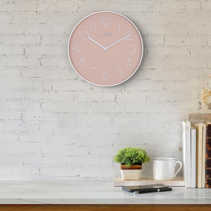 Lifestyle image of the 404-3831P 12-inch Paxton Wall Clock - Peach, with Silent Sweep, hanging on a white-washed brick wall above a marble countertop with books, a plant, and a phone. The clock features a flush-mount peach frame, a matching color dial, white raised numbers, and white metal hour, minute, and second hands.