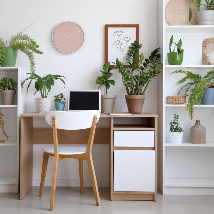 Lifestyle image of the 404-3831P 12-inch Paxton Wall Clock - Peach, with Silent Sweep, shown hanging on a wall in a home office with a desk, bookshelves, and plants. The clock features a flush-mount peach frame, a matching color dial, white raised numbers, and white metal hour, minute, and second hands.