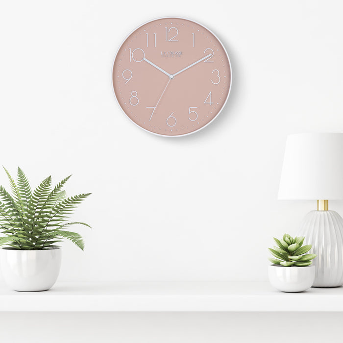 Lifestyle image of the 404-3831P 12-inch Paxton Wall Clock - Peach, with Silent Sweep, hanging on a white wall above a white shelf with a white lamp and plants in white vases. The clock features a flush-mount peach frame, a matching color dial, white raised numbers, and white metal hour, minute, and second hands.