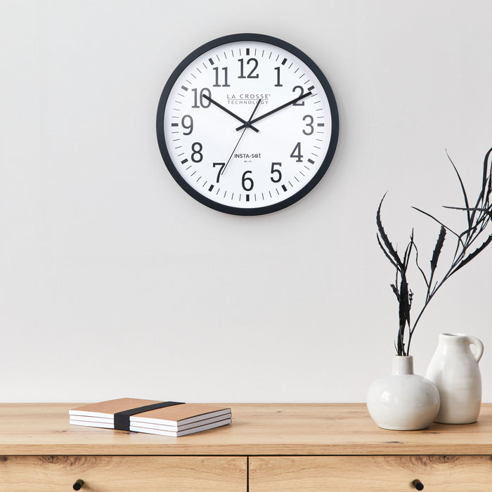 Lifestyle image of the 404-3836 Insta-Set Wi-Fi Wall Clock for iOS users. The clock features a black flush-mount frame, a white dial with black numbers, and black hands, and is shown hanging on a light gray wall above a Scandinavian design side table in light wood with a white vase and a white jug with black sticks beside a stack of notebooks.
