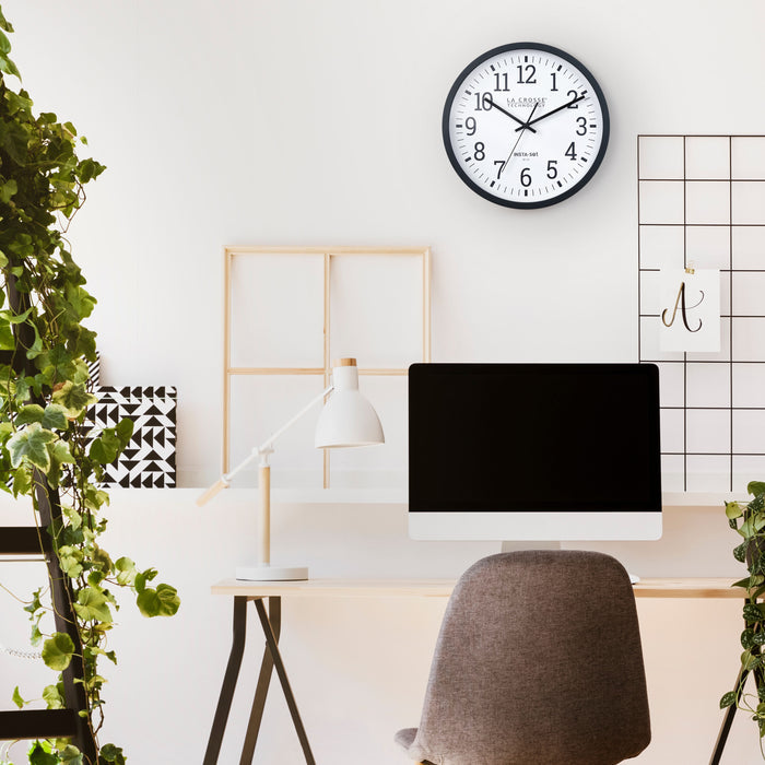 Lifestyle of a modern work station with a computer monitor, lamp, chair, and plants with the 404-3836 Insta-Set Wi-Fi Wall Clock for iOS users hanging on the wall. The clock features a black flush-mount frame and a white dial with black numbers and black hands.