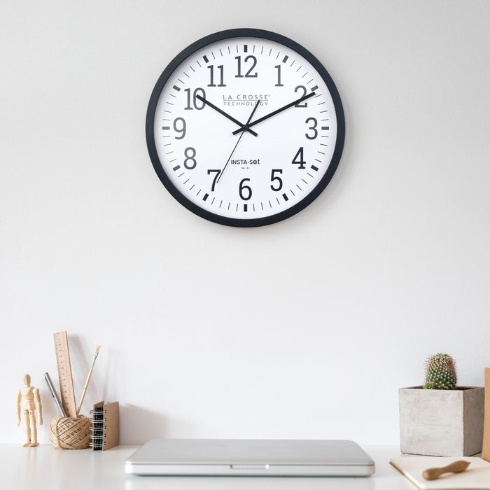 Lifestyle of the 404-3836 Insta-Set Wi-Fi Wall Clock for iOS users. The clock features a black flush mount frame, white dial with black numbers, and black hands hanging on a light gray wall over a white desk with a closed laptop, a cactus, a figurine, and other desk accessories