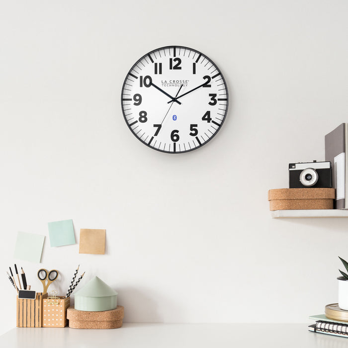Lifestyle image of the 404-4536 14.2-inch Insta-Set Wall Clock with Bluetooth® Technology shown hanging on a white wall above a white desk with desk accessories and a shelf holding books, a cork box, and a camera. The clock features a white dial with bold black numbers and markings, a black flush-mount frame, and black hands.