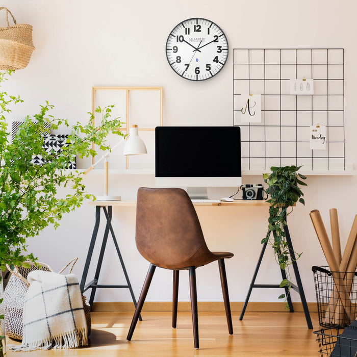 Lifestyle image of the 404-4536 14.2-inch Insta-Set Wall Clock with Bluetooth® Technology, shown hanging on a tan wall above a natural-color desk with a lamp, computer, plants, and a brown leather desk chair. The clock features a white dial with bold black numbers and markings, a black flush-mount frame, and black hands.