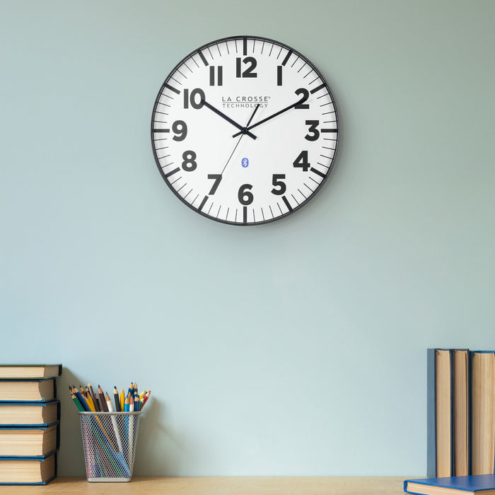 Lifestyle image of the 404-4536 14.2-inch Insta-Set Wall Clock with Bluetooth® Technology, shown hanging on a light seafoam green wall above a natural-color desk with books and a pencil container. The clock features a white dial with bold black numbers and markings, a black flush-mount frame, and black hands.