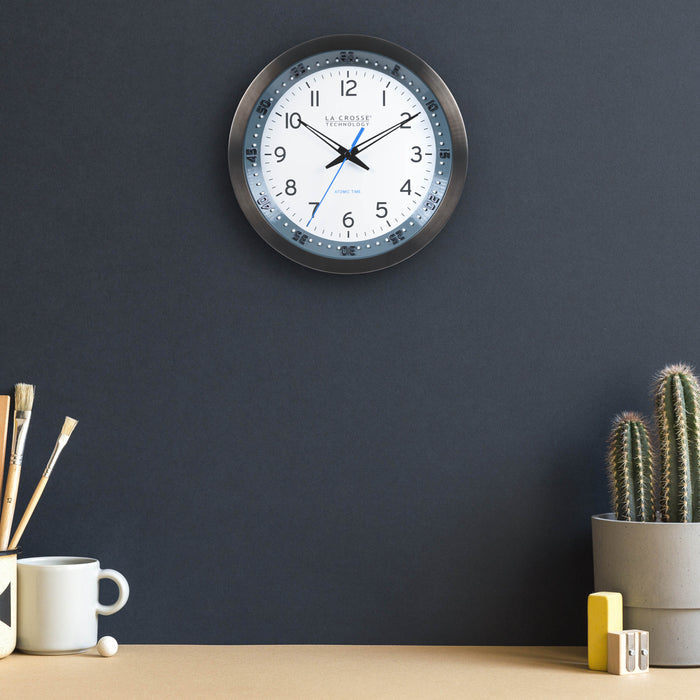 Lifestyle image of the 404-54667 10-inch Atomic Metal Wall Clock with Blue Chapter Ring hanging on a navy wall over a light wood color desk with a cup, paint brushes, and a cactus. The clock features a black stainless steel frame with a blue chapter ring and raised seconds indicators, surrounding a white dial with black numbers, metal hands, and a blue accent second hand.