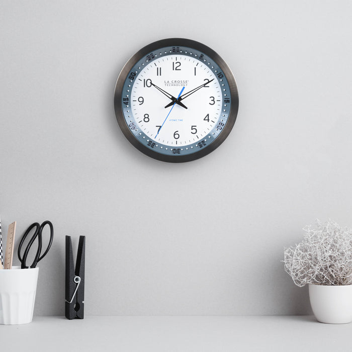 Lifestyle image of the 404-54667 10-inch Atomic Metal Wall Clock with Blue Chapter Ring hanging on a gray wall over a gray desk with a white pencil cup a letter clip and a plant. The clock features a black stainless steel frame with a blue chapter ring and raised seconds indicators, surrounding a white dial with black numbers, metal hands, and a blue accent second hand.