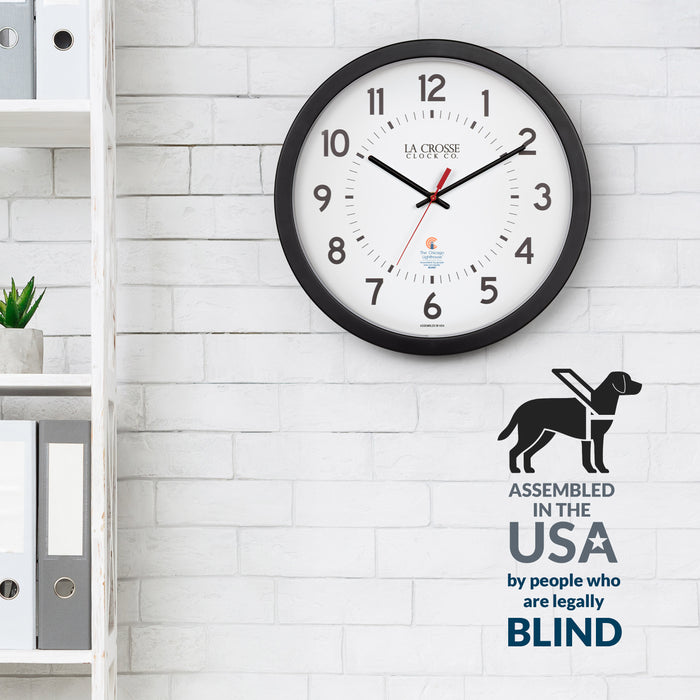 Lifestyle image of the 404-5537 14.5-inch Wall Clock hanging on a white brickwall next to a white bookshelf with binders and plants. The clock features a black flush mount frame, black bold numbers, black hour and minute hands, and a red second hand with two logos on the clock face, La Crosse Clock Co. and Chicago Lighthouse industries. The image features the CLI badge stating "assembled in the USA by people who are legally blind"