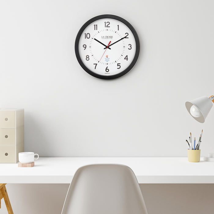 Lifestyle image of the 404-5537 14.5-inch Wall Clock hanging on a wall over a white desk, white chair, and lamp with desk accessories. The clock features a black flush mount frame, black bold numbers, black hour and minute hands, and a red second hand with two logos on the clock face, La Crosse Clock Co. and Chicago Lighthouse industries.