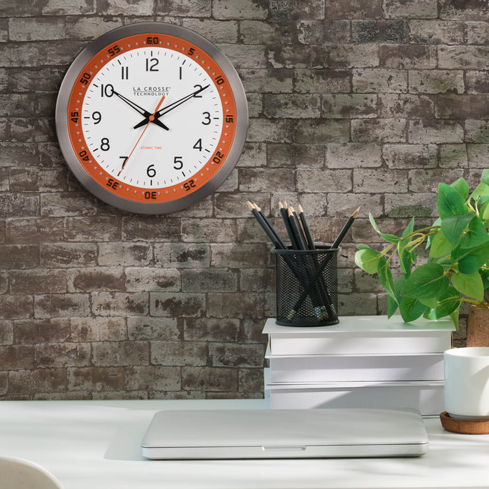 Lifestyle image of the 404-87696 10-inch Atomic Metal Wall Clock - Chapter Ring: Orange shown hanging on a brick wall over a desk with a computer and desk accessories. The clock features a white dial, an orange chapter ring with seconds indicator, black hands, an orange second hand, and a stainless steel flush-mount frame.