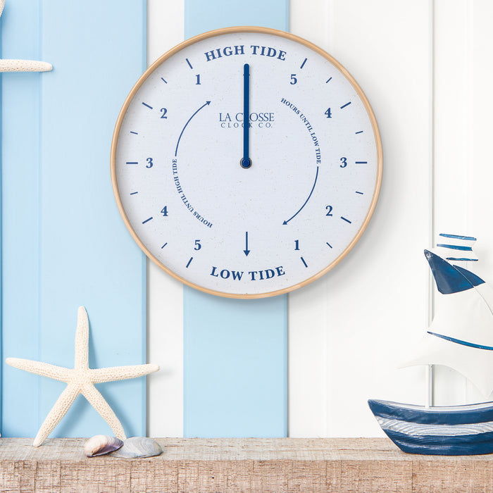 Lifestyle image of the 425-3831 11.8-inch Tide Wall Clock shown hanging on a blue and white slate wall above a rustic wood shelf with nautical accents, a ship, starfish, and shells. The clock features a light wood frame, a white and tan speckled dial, blue numbers, markings, and a blue metal hand.