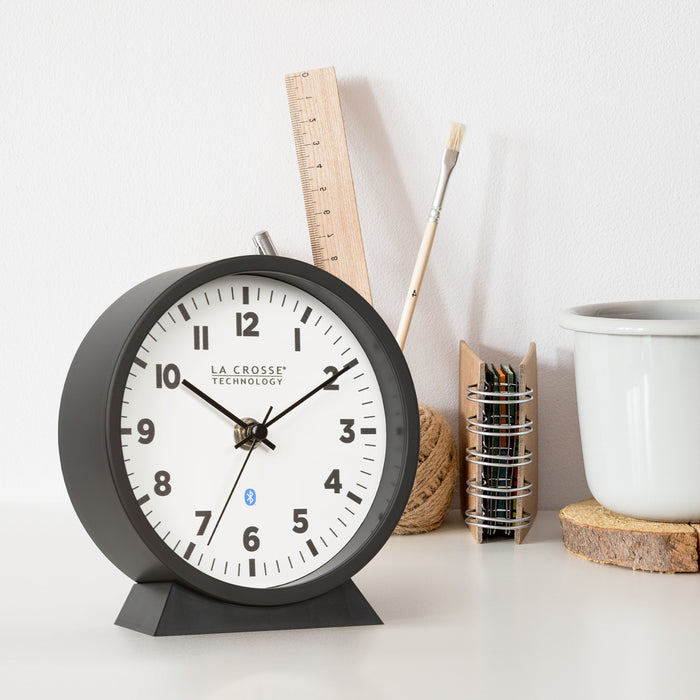 Right-facing lifestyle image of the 437-3816 Insta-Set Table/Wall Clock with Bluetooth® Technology shown sitting on a desk with a coffee cup, pencil holder, and notebook. The clock features a white dial with bold black numerals, black metal hands, and a black frame with removable base for tabletop or wall hanging placement.