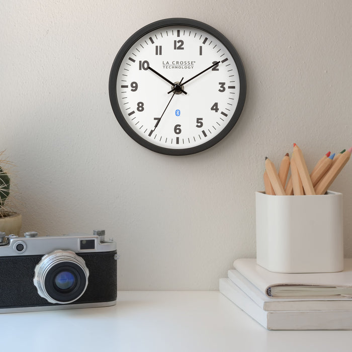 Lifestyle image of the 437-3816 Insta-Set Table/Wall Clock with Bluetooth® Technology shown hanging on a white wall over a desk with books, a pencil cup, and a vintage camera. The clock features a white dial with bold black numerals, black metal hands, and a black frame with removable base for wall hanging placement.