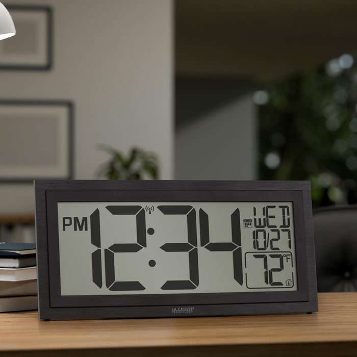 Lifestyle image of the 515-1419v2 Atomic Digital Wall Clock with Time Advance shown on a wood desk in an office setting. The digital wall clock features a dual-textured black frame, easy-to-read 4.49-inch time digits, indoor temperature reading of 72 degrees Fahrenheit, and a calendar.