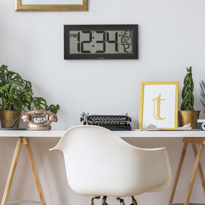 Lifestyle image of the 515-1419v2 Atomic Digital Wall Clock with Time Advance shown hanging on the wall over a white desk in a home office setting. The digital wall clock features a dual-textured black frame, easy-to-read 4.49-inch time digits, indoor temperature reading of 72 degrees Fahrenheit, and a calendar.