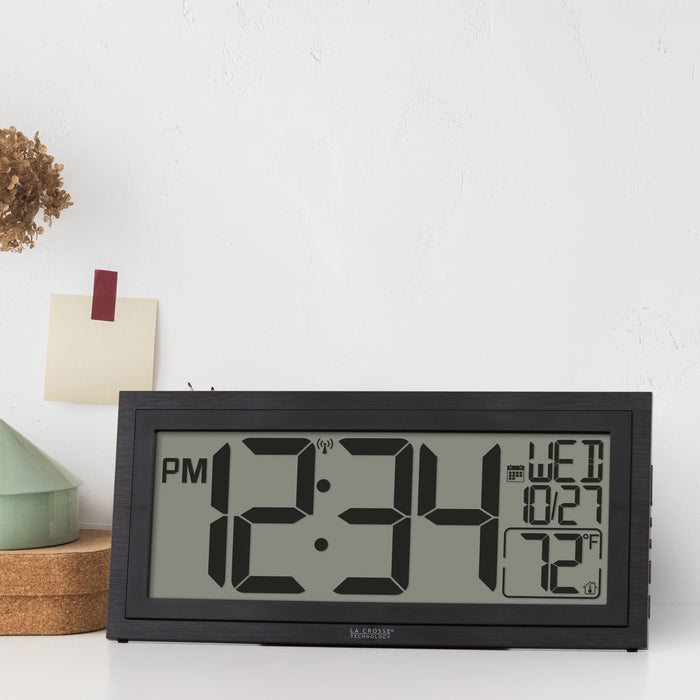 Lifestyle image of the 515-1419v2 Atomic Digital Wall Clock with Time Advance shown on a white desk. The digital wall clock features a dual-textured black frame, easy-to-read 4.49-inch time digits, indoor temperature reading of 72 degrees Fahrenheit, and a calendar.
