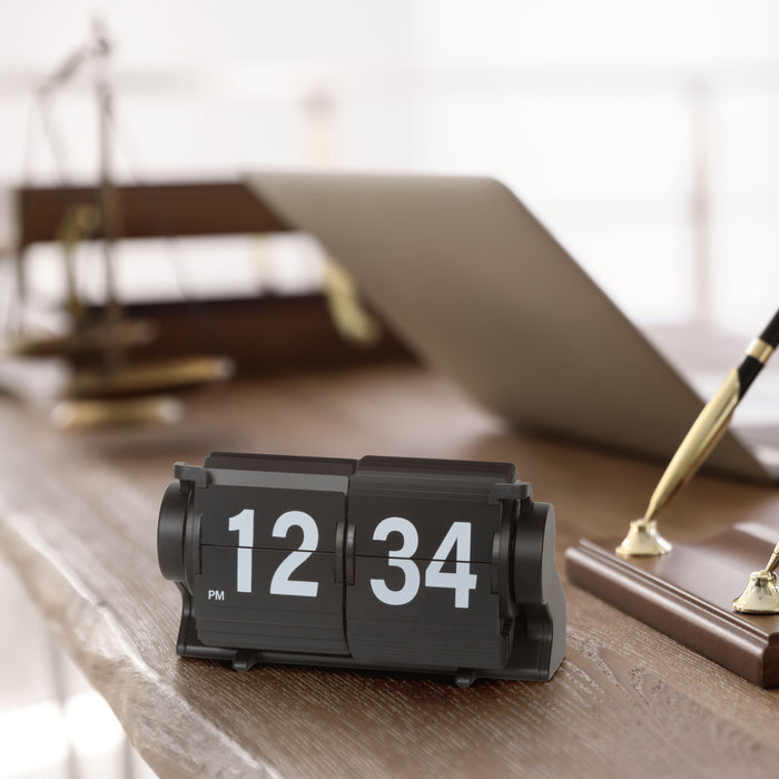 Lifestyle image of the 536-527B Rollins Mini Mechanical Flip Clock – Black, shown sitting on a dark wood live-edge desk next to a wood base pen set in gold and black with a computer in the background. The clock features black hour and minute flip cards with white numbers in a black case.