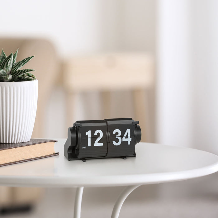 Lifestyle image of the 536-527B Rollins Mini Mechanical Flip Clock - Black, shown sitting on a white side table with a book and a fluted pot with a plant and a set of chairs in the background. The clock features black hour and minute flip cards with white numbers in a black case.