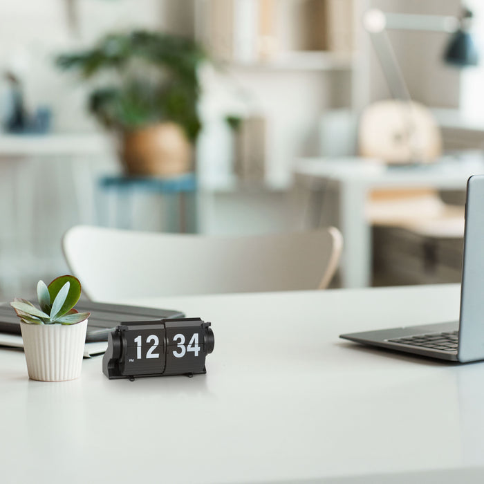Lifestyle image of the 536-527B Rollins Mini Mechanical Flip Clock - Black  shown sitting on a white desk next to a white potted plant with an office setting in the background. The clock features black hour and minute flip cards with white numbers in a black case.