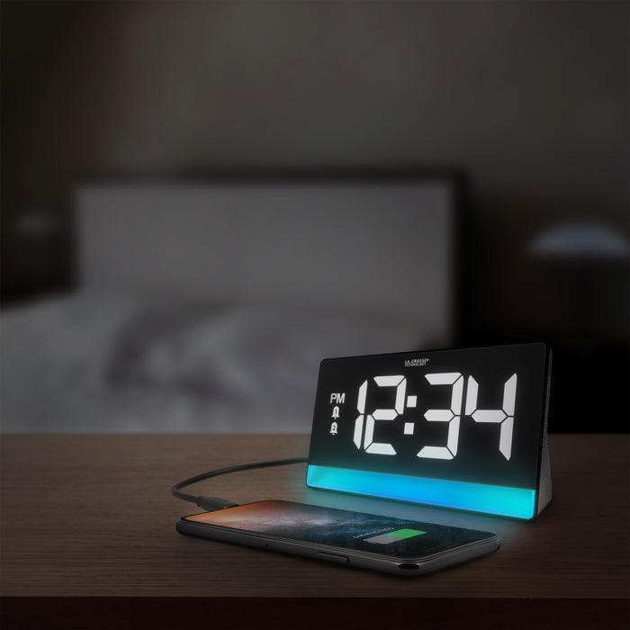 Lifestyle image of the 617-149 Digital Alarm with Color Changing Glow Light shown on a nightstand with a bed in the background in a darkened bedroom. The alarm clock features a white case, a color-changing bar as the base, and a black display with white digits.